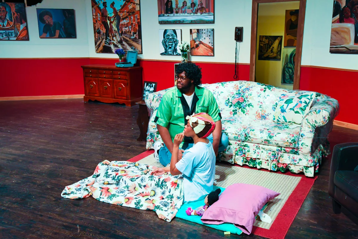 Two performers in a scene beside a floral sofa on an artwork-lined stage.